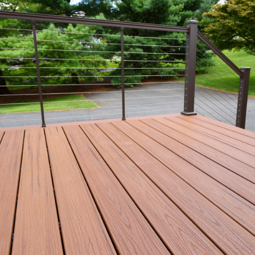 Light brown composite decking with chocolate brown railing; a driveway lined with pine trees visible through the railing.