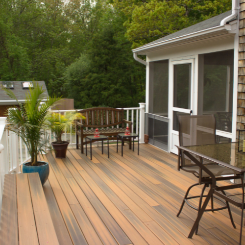 Tones of brown composite decking surrounded by white railing, featuring patio furniture including a wooden bench, a glass table with brown rim, and chairs. Trees are visible, and a house with white trim, doors, and a screened-in room is partially seen in the background.